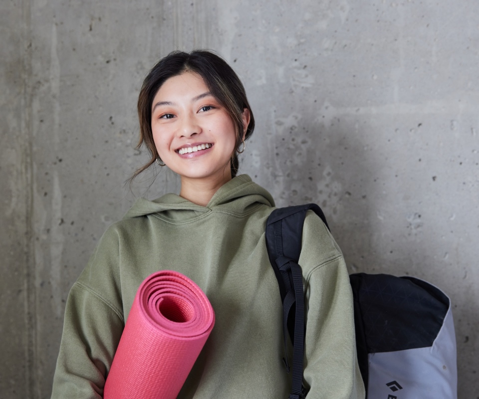 Young woman smiling while holding a pink yoga mat, dressed in a casual green hoodie and carrying a backpack.