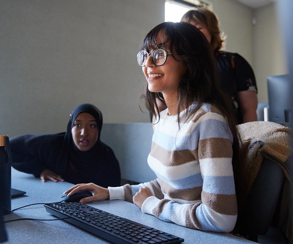 Two women engaged in a computer-based learning environment, one actively working at a desk while the other observes with interest.