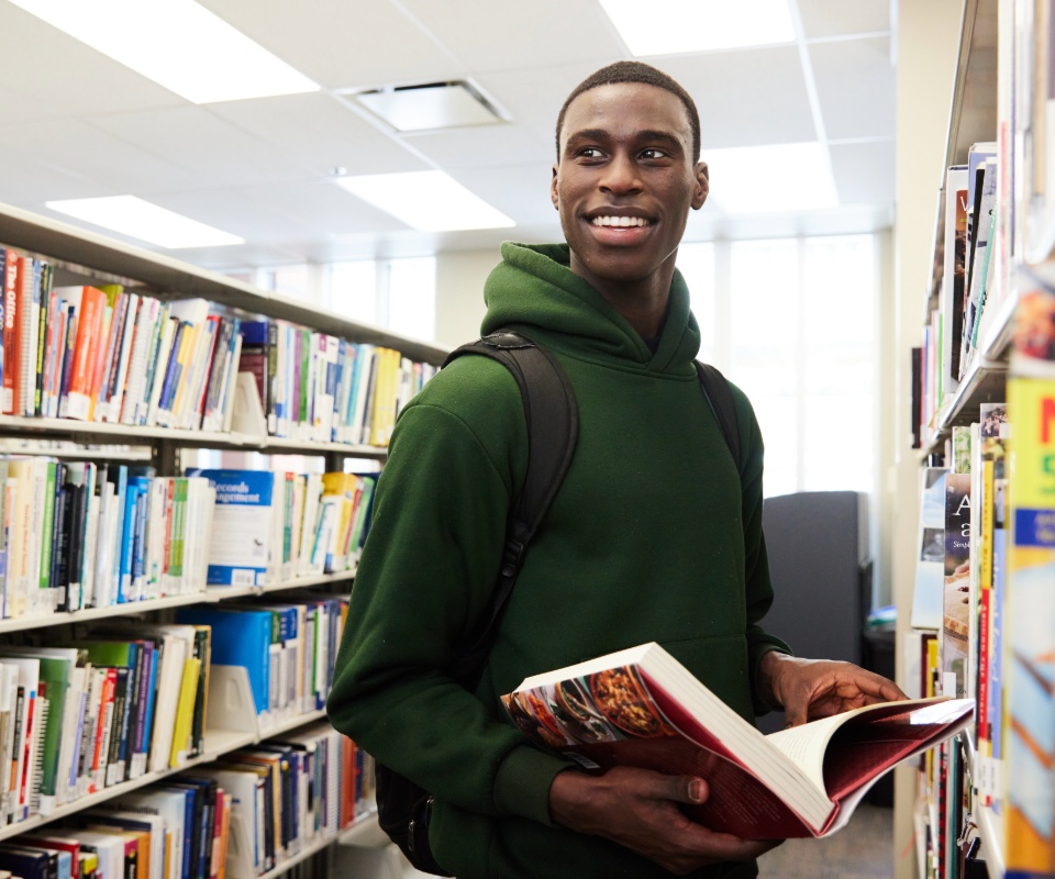 A smiling student holding an open book while standing in a library filled with shelves of books.