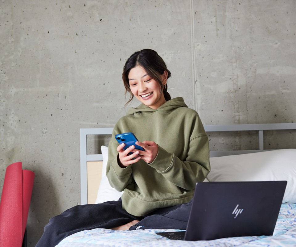 Young woman sitting on a bed, smiling while using her smartphone, with a laptop beside her.