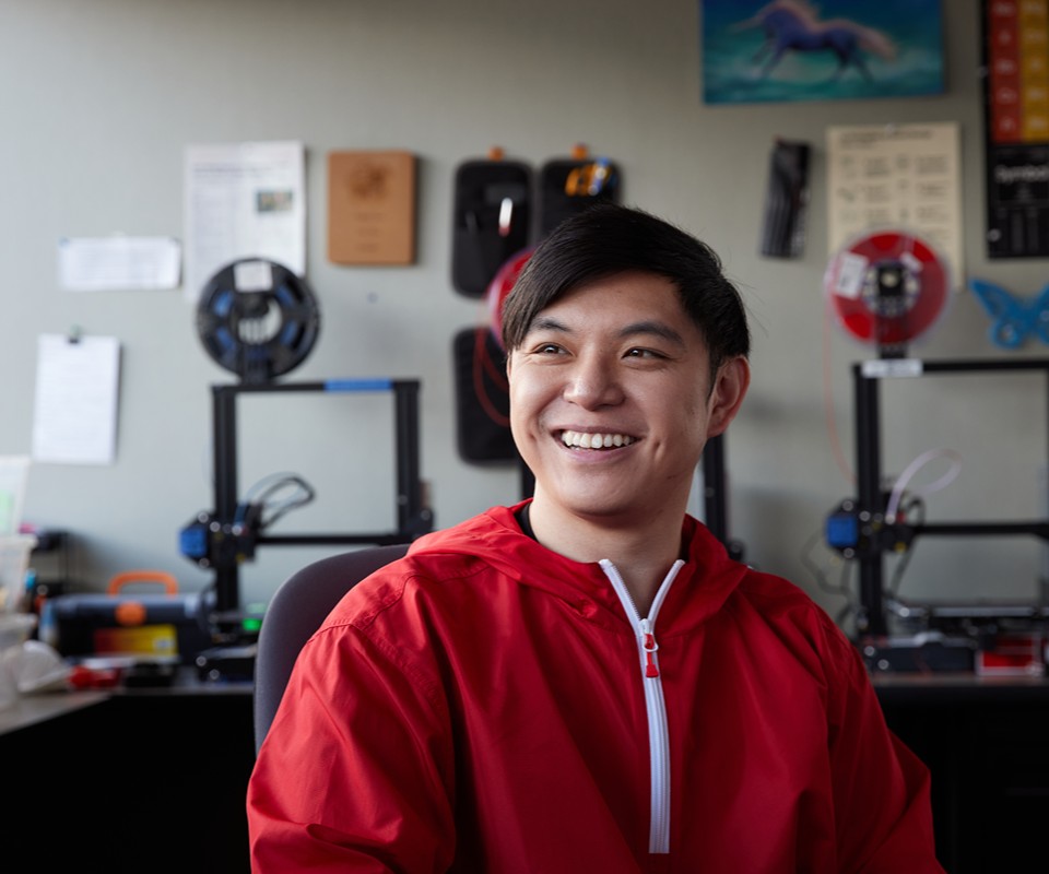 A young man wearing a red jacket smiles while seated in a workshop with 3D printers in the background.