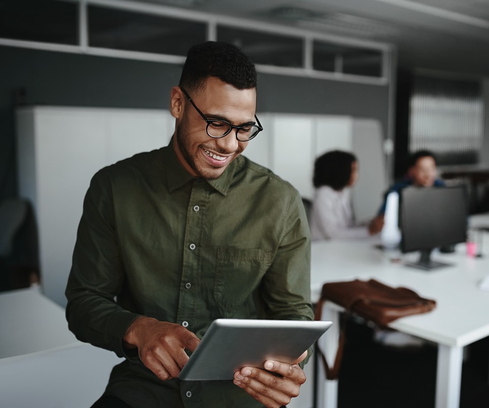 A smiling man in glasses reads a tablet while seated in a modern office environment, with colleagues visible in the background.
