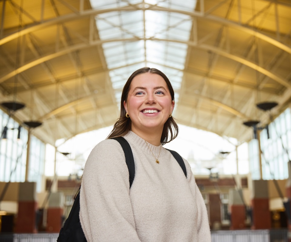 A smiling young woman wearing a beige sweater stands in an airy, modern building with a high ceiling and large windows.