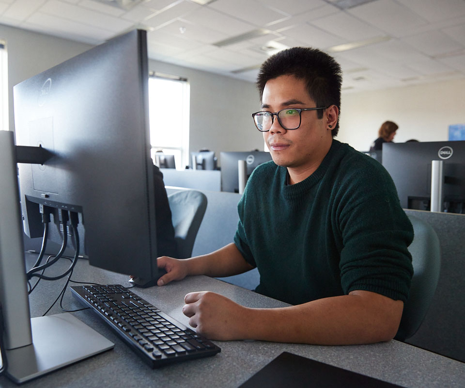A student working on a computer in a classroom setting, focused on the screen.