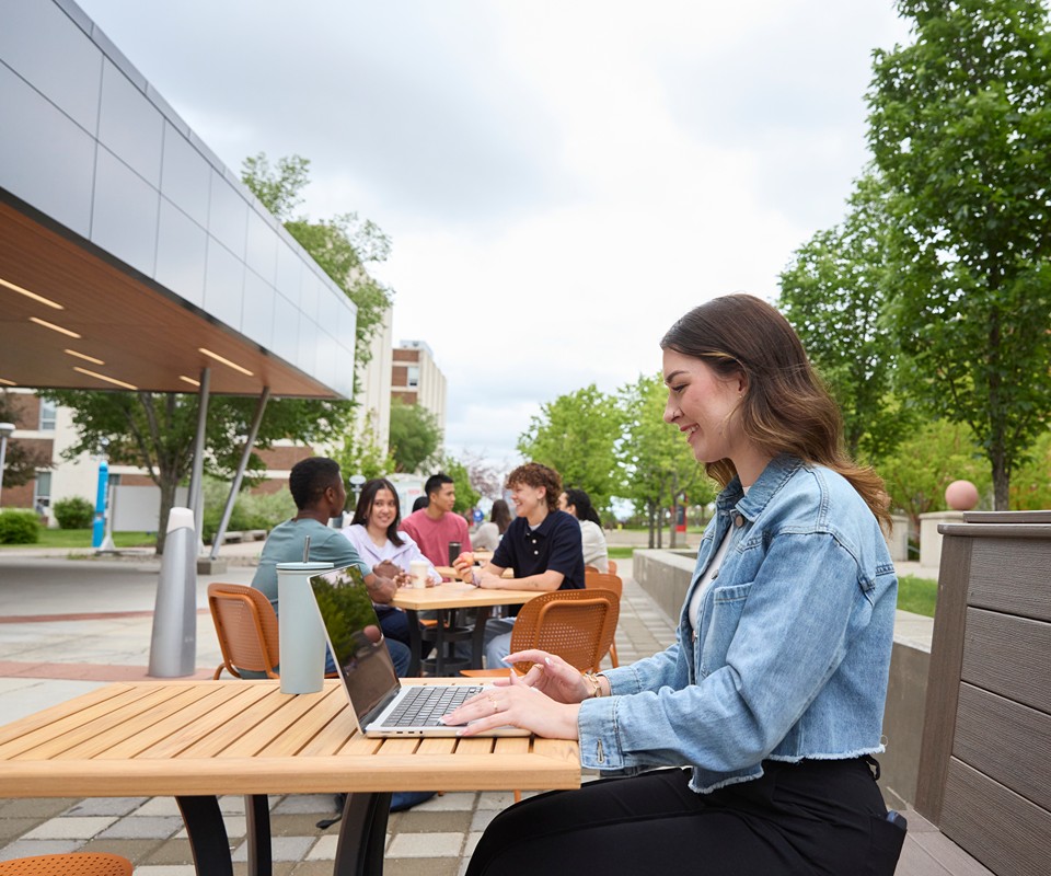 Student sits outside John Ware on SAIT campus, typing on a laptop and smiling.
