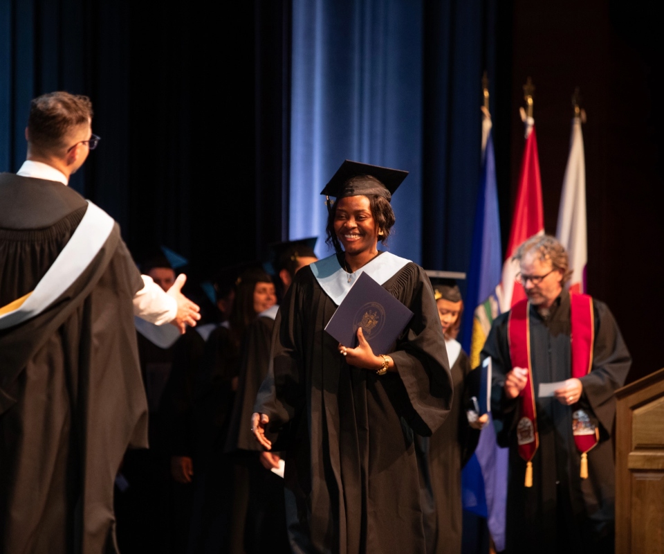 A smiling graduate receiving a diploma on stage during a graduation ceremony, with faculty and flags in the background.