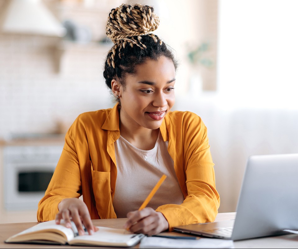 A young woman with braided hair wearing a yellow shirt is taking notes while working on a laptop at a wooden desk.