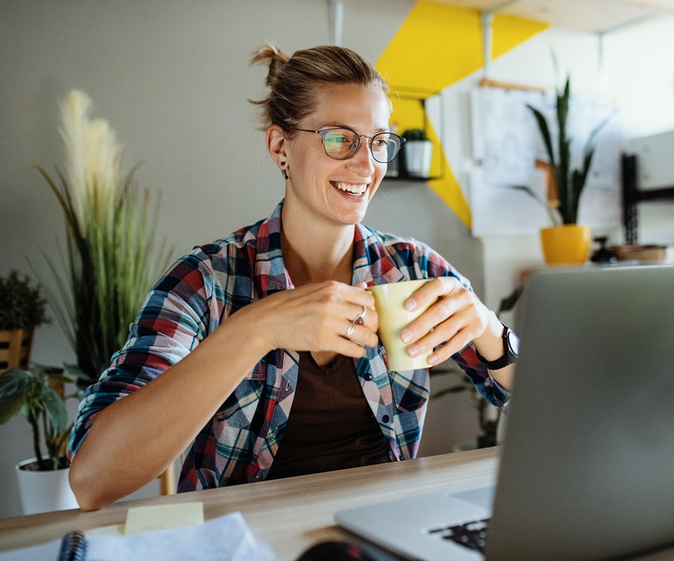 A student smiles while working on a laptop, holding a cup in a cozy office space filled with plants.