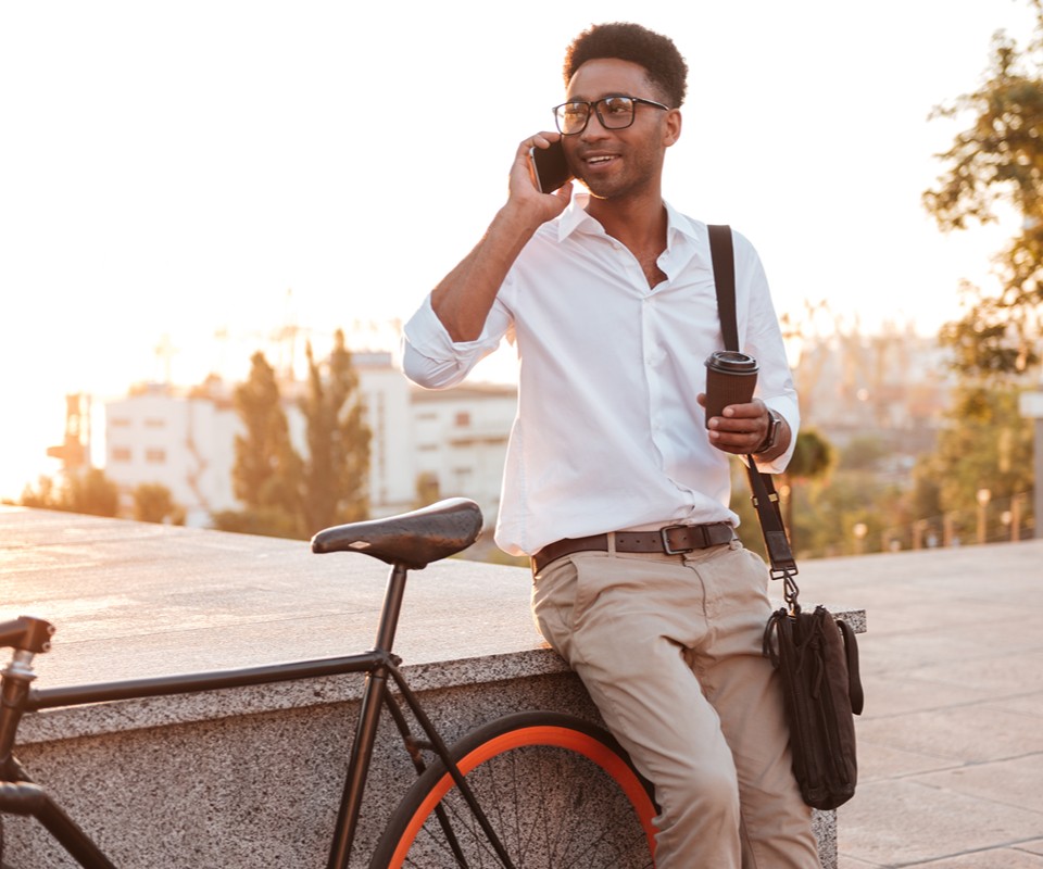 Young man in glasses talking on the phone, standing next to a bicycle while holding a coffee cup.