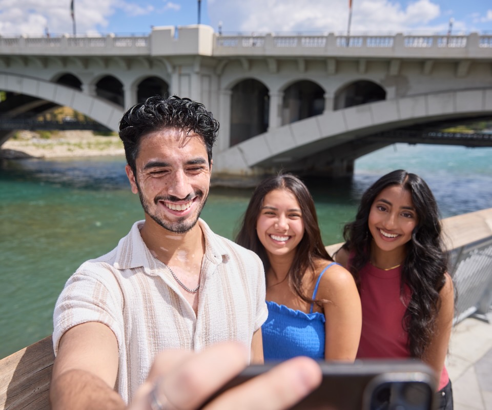 A group of three students take a selfie by a bridge on a sunny day.