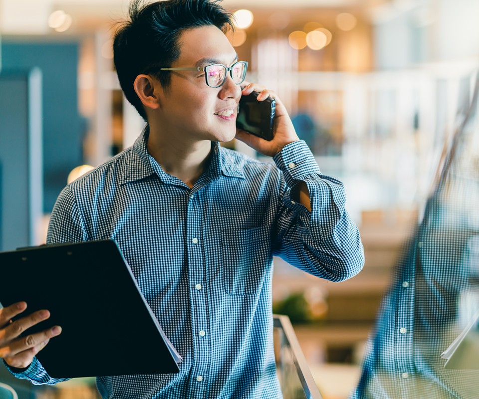 A professional man in a checkered shirt talking on his phone while holding a clipboard, smiling pleasantly.