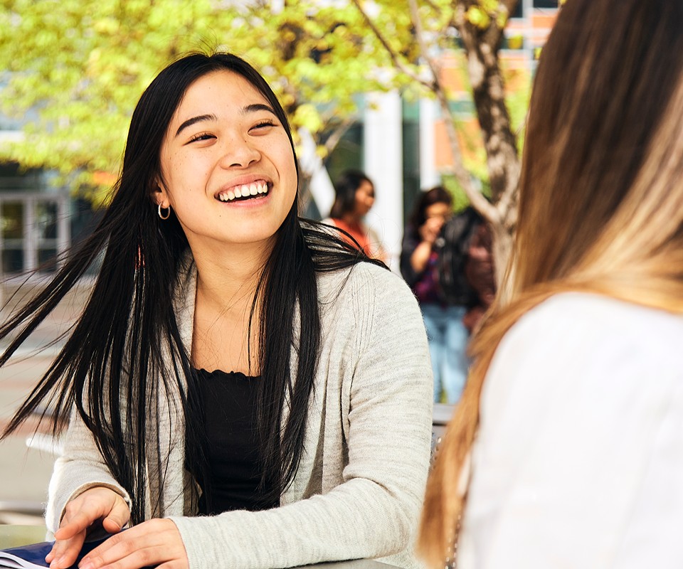 A smiling young woman engaged in conversation outdoors, surrounded by greenery and fellow students in the background.