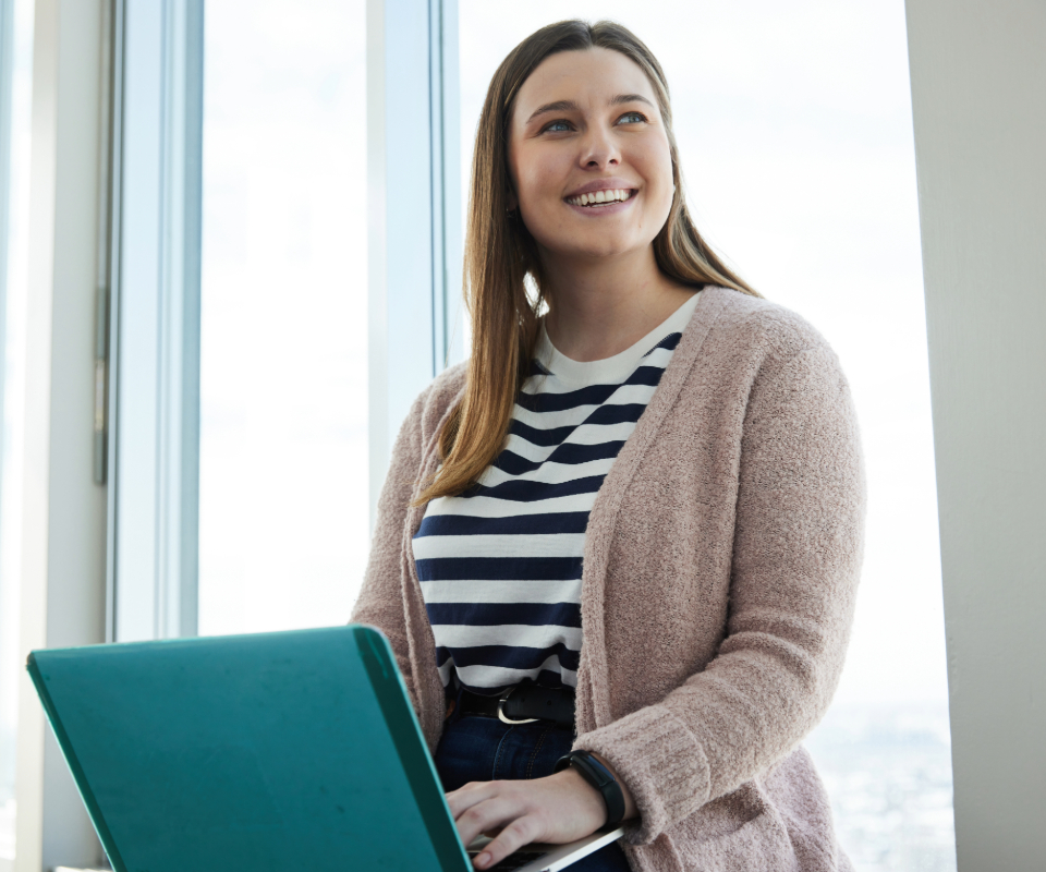 A young woman with long brown hair is seated at a window, smiling while working on a laptop. She is wearing a striped shirt and a cozy pink cardigan.