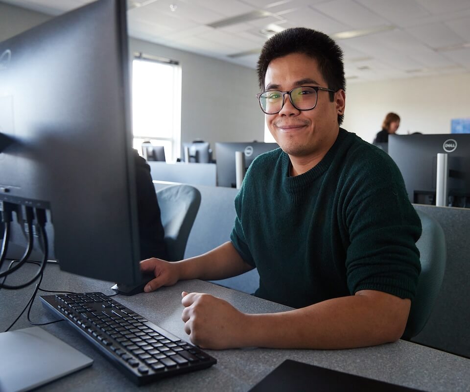 A student sitting at a desktop computer smiles at the camera