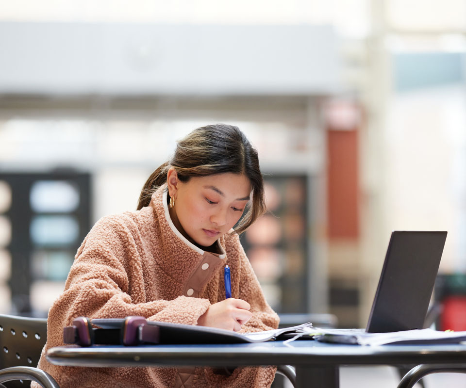 A student focused on her work, writing notes while seated at a table with a laptop and papers.