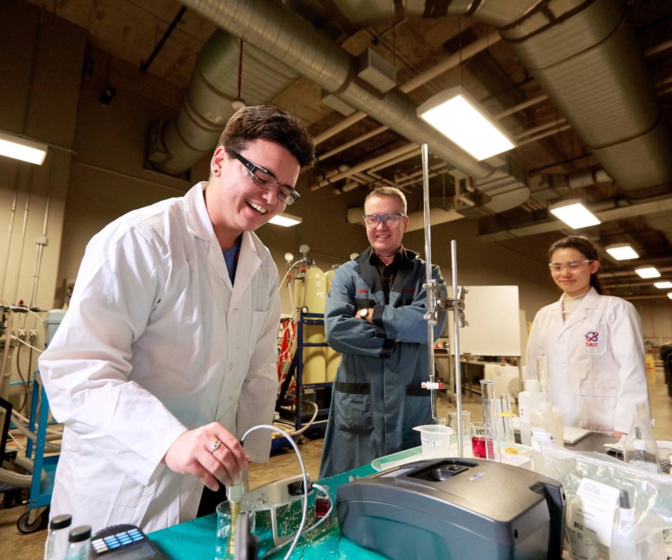 Two students and a teacher are engaged in a research activity, smiling and wearing lab coats.