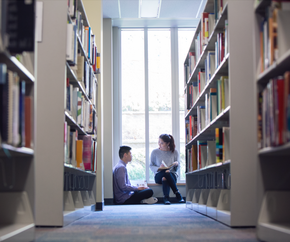 Two students sit on the floor of a library aisle, engaged in discussion while surrounded by bookshelves.