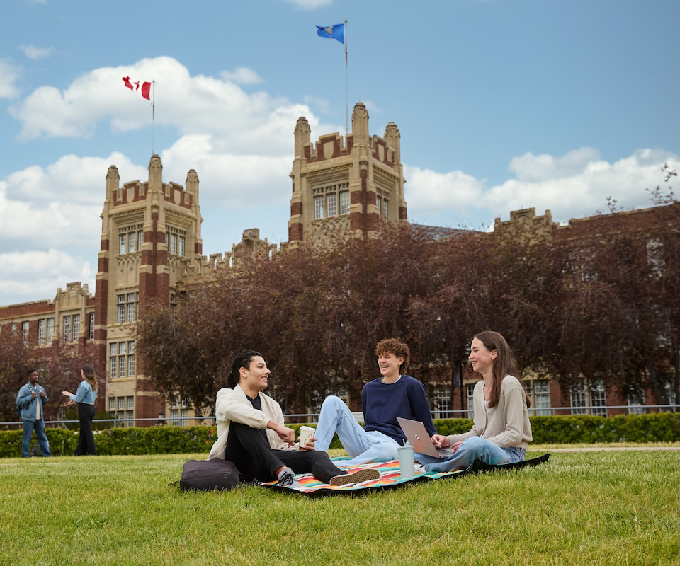 Three students sit on a blanket on a grassy area, engaged in work and conversation, with a historic college building and flags in the background.