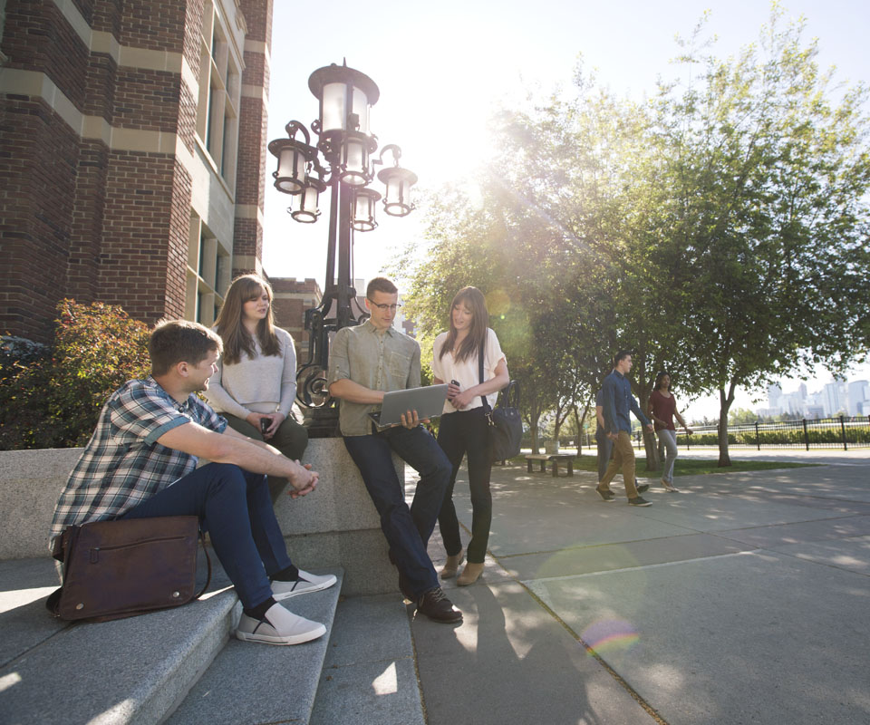 A group of four students engaged in a discussion on the steps of Heritage Hall, with one person using a laptop while sitting on steps next to a lamp post. Bright sunlight filters through trees in the background.