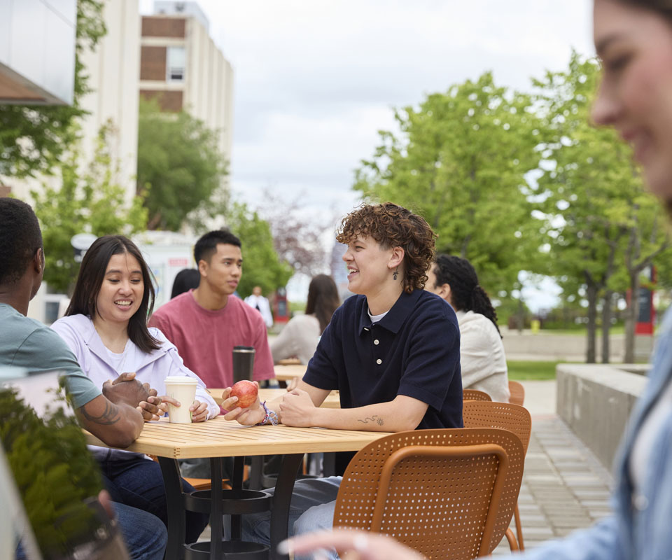 A diverse group of students enjoying a casual conversation at an outdoor table on campus.