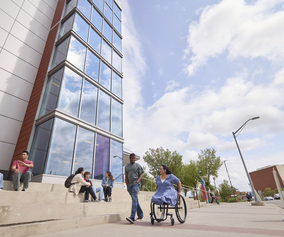 A ground of students gathered outside the Begin Tower residence during the summer. Some students sit on concrete steps, some are walking and one woman is in a wheelchair.