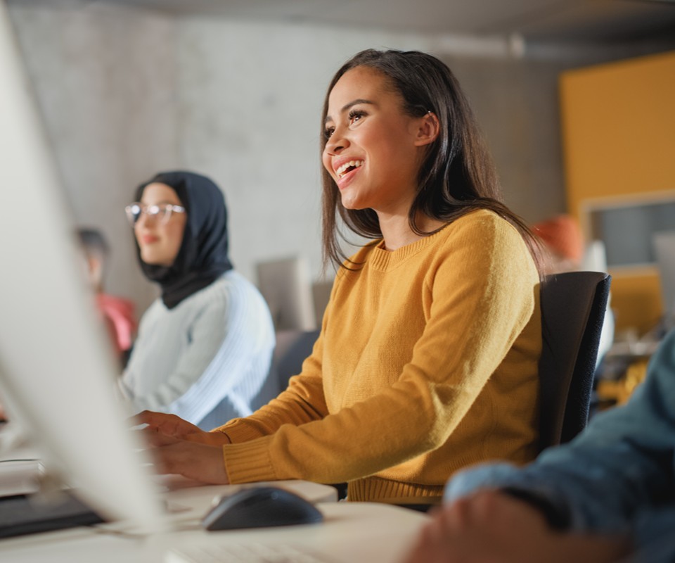 A young woman in a yellow sweater smiles while working at a computer in a collaborative learning environment.