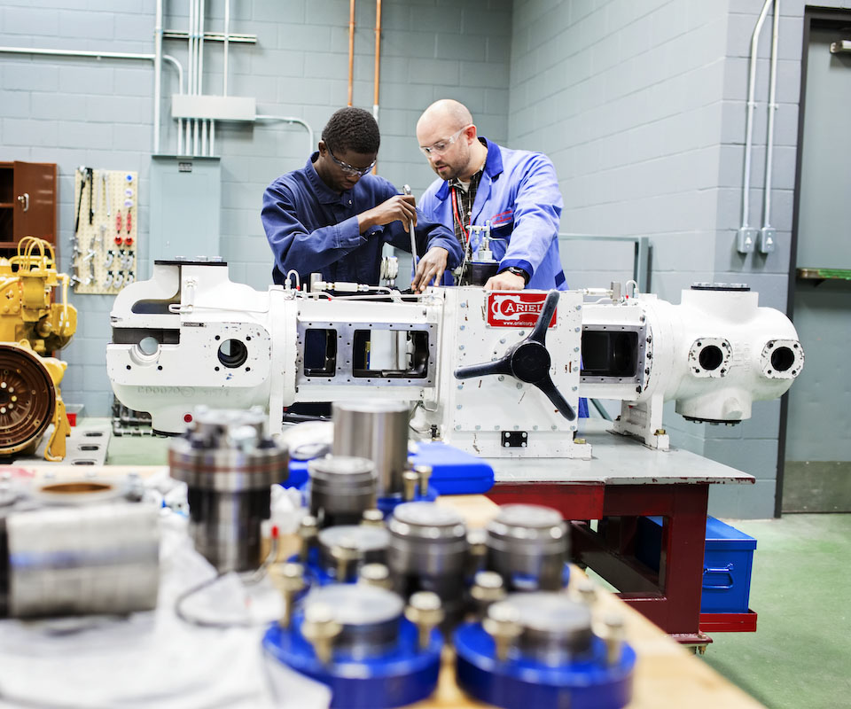 Two apprentices work together on a large mechanical assembly in a workshop, surrounded by various tools and machinery components.