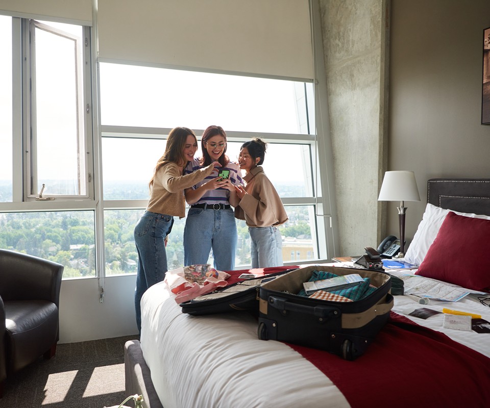 Three friends joyfully looking at a smartphone in a modern hotel room with large windows and a suitcase on the bed.