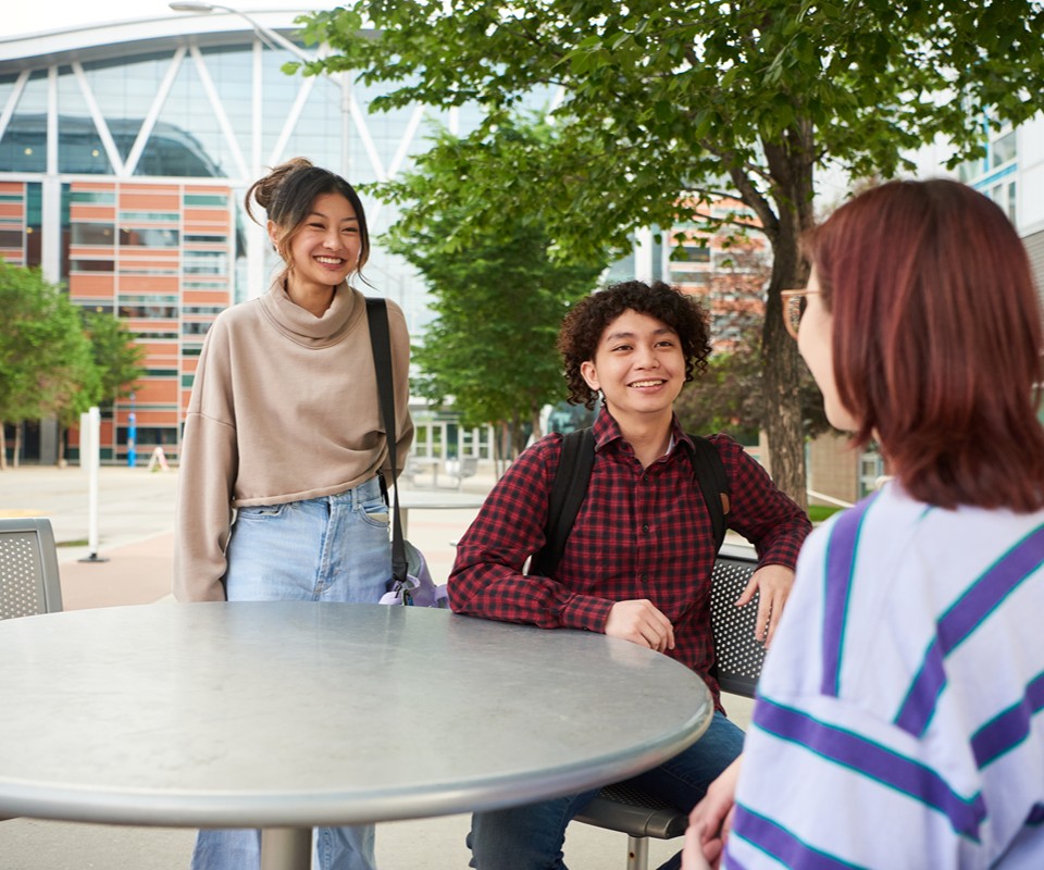 Three students engaged in conversation at an outdoor table on a college campus. The atmosphere appears friendly and social.