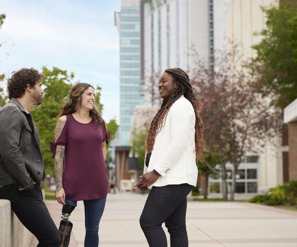 Three diverse people engage in a friendly conversation outdoors, set against a backdrop of the modern Begin Tower residence and greenery.