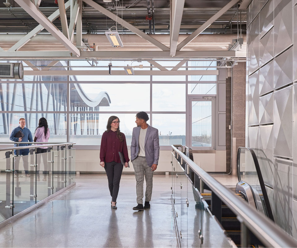 Two professionals walk side by side in a modern, well-lit hallway with large windows within the Johnson-Cobbe Energy Centre. The scene conveys a collaborative and dynamic environment.