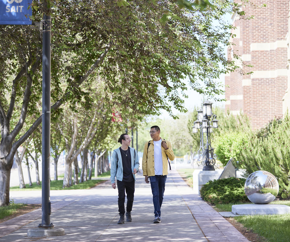 Two men walking side by side on a tree-lined pathway, engaging in conversation.