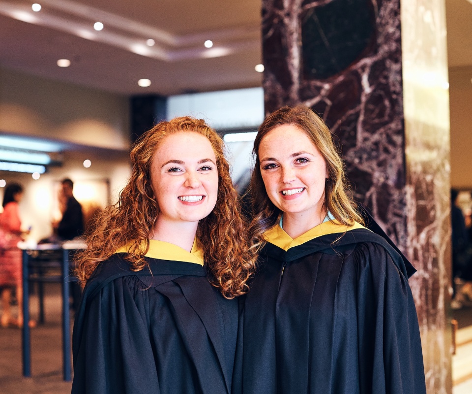 Two female graduates wearing black gowns and yellow hoods smile together in a celebratory setting.