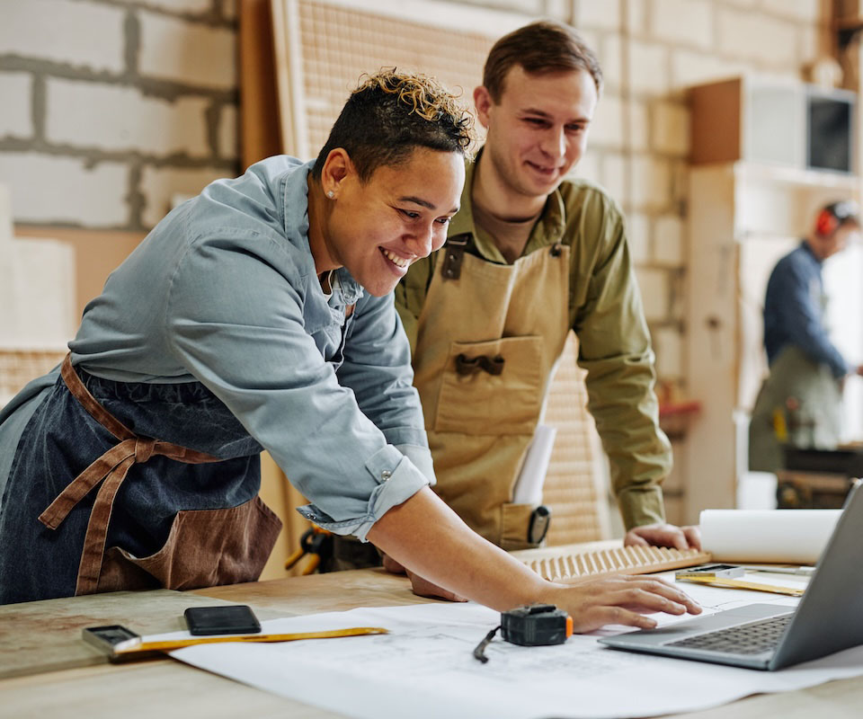 Two smiling apprentices collaborating over a laptop in a workshop, showcasing teamwork and creativity in a craft environment.