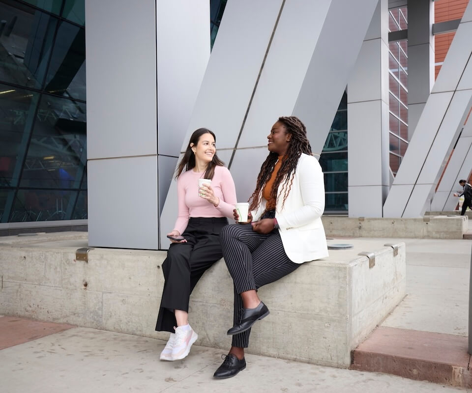 Two people sitting outside on a concrete slab having a hot drink and talking