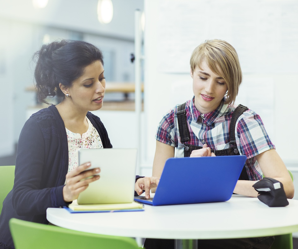 Two women have a discussion while looking at information on a laptop and tablet.