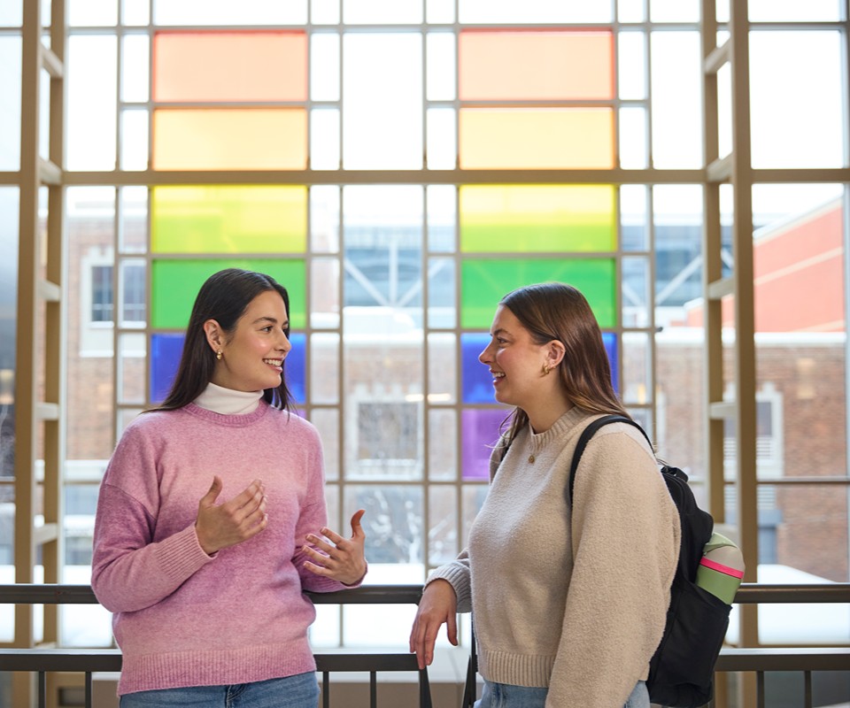 Two women engage in conversation in front of a colorful rainbow window display.