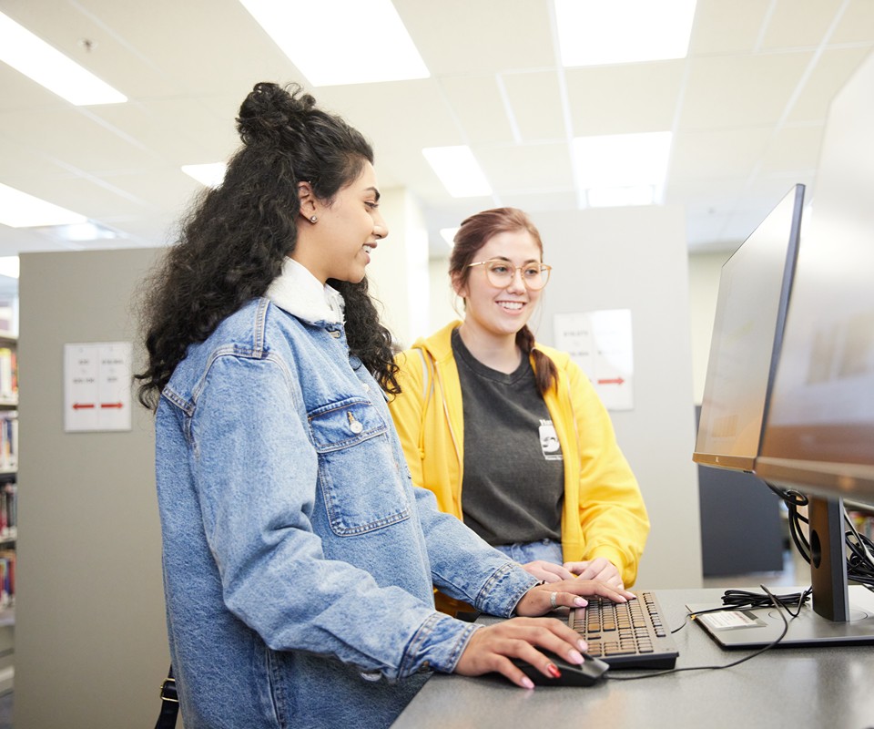 Two students collaborating at a computer in a library, one wearing a denim jacket and the other in a yellow hoodie.