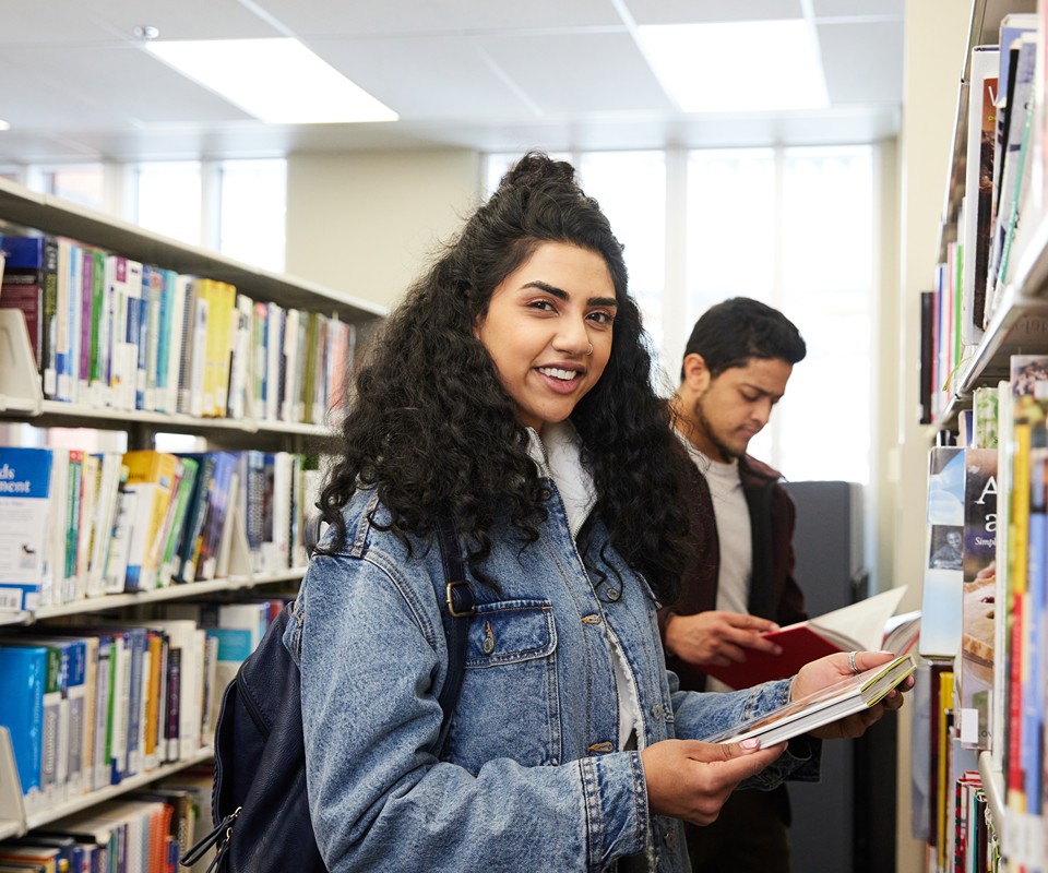 Young woman in a denim jacket smiles at the camera while browsing books in a library, with a man reading in the background.