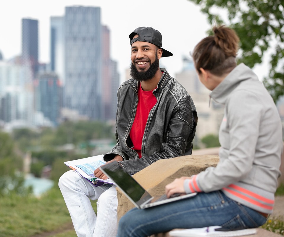 A smiling man in a leather jacket sits outdoors with a woman working on a laptop, set against a city skyline.