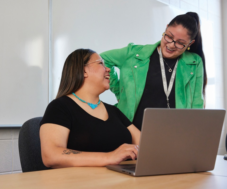 Instructor laughing a student in the classroom, with a laptop nearby.