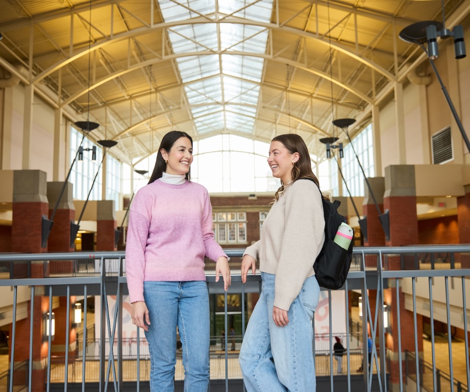 Two students standing in a campus atrium – talking beside a railing beneath a large skylight and open gathering space.
