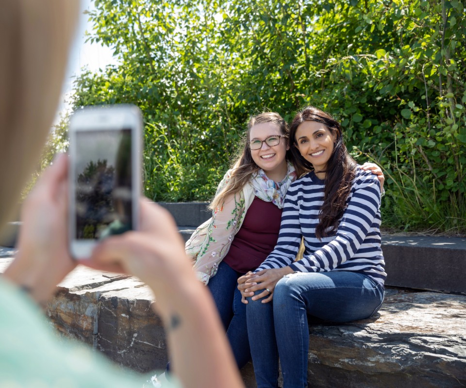 Two women sitting on a rock, smiling together while one takes a photo of them with a smartphone amidst greenery.