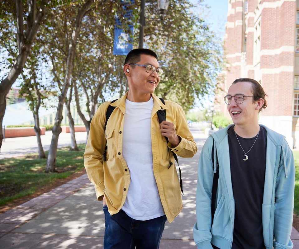 Two students walking outdoors on a campus pathway, engaged in conversation and smiling. The setting features trees and buildings in the background.