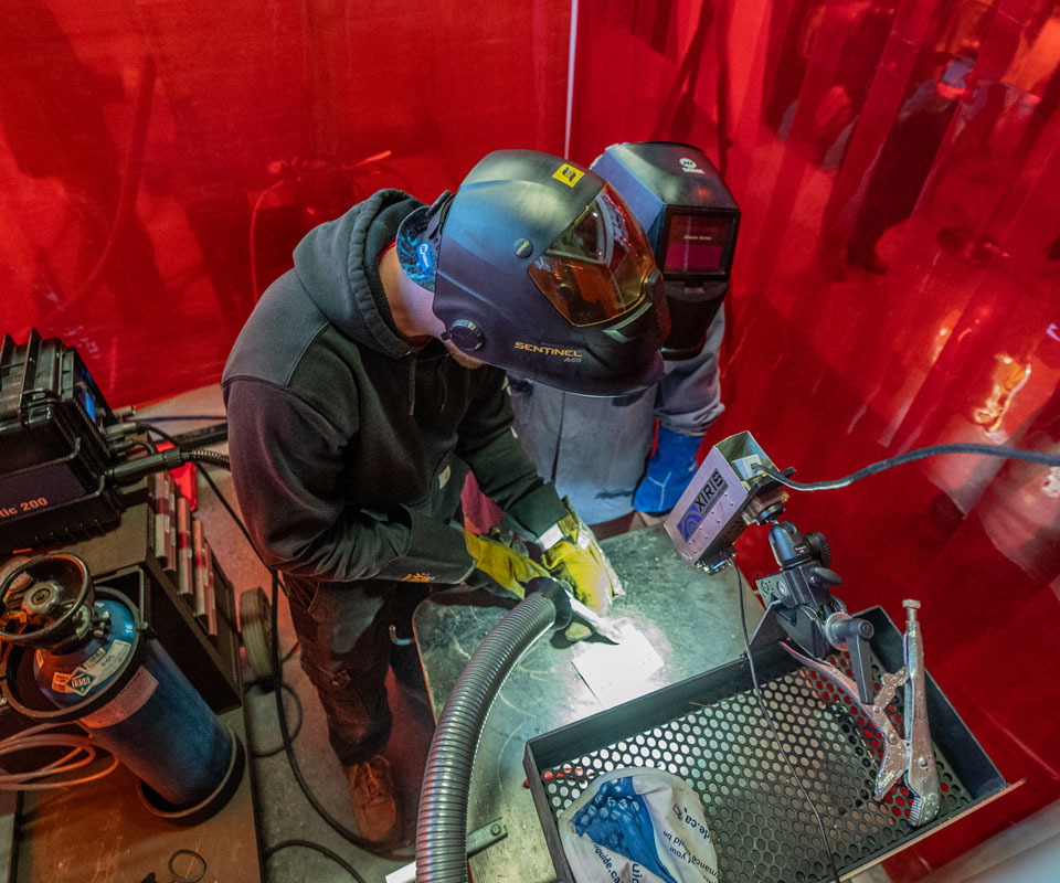 A welding student using specialized equipment in a red-tinted work environment. The image highlights the welding process and safety gear.