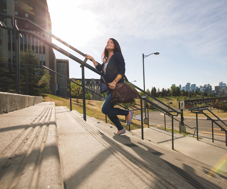 A woman ascends outdoor stairs, carrying a brown bag, with a city skyline in the background. Sunlight illuminates the scene, creating a warm and vibrant atmosphere.
