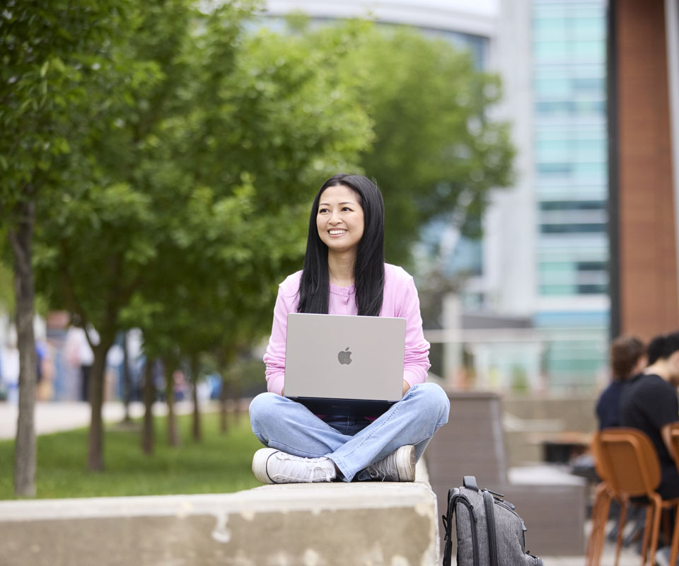 A young woman sitting cross-legged outdoors with a laptop, smiling while surrounded by greenery and fellow students.