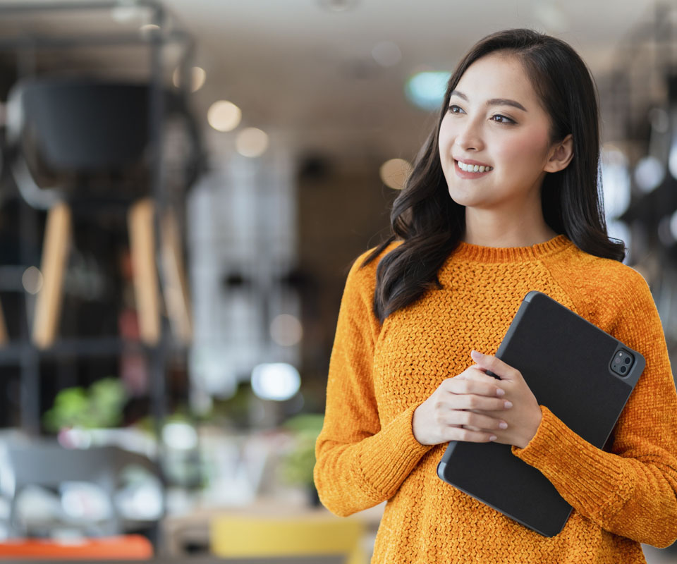 A smiling woman in an orange sweater holding a tablet in a modern café setting.