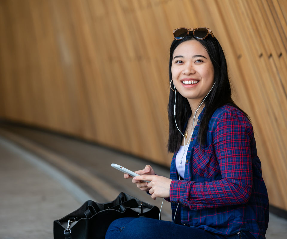 A smiling young woman wearing headphones holds a smartphone while sitting against a wooden wall.