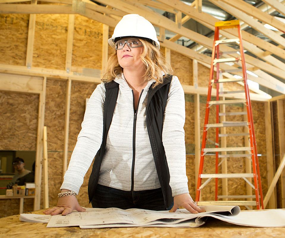 A woman in a hard hat and glasses reviews construction plans on a table, with a ladder and wooden framework in the background.